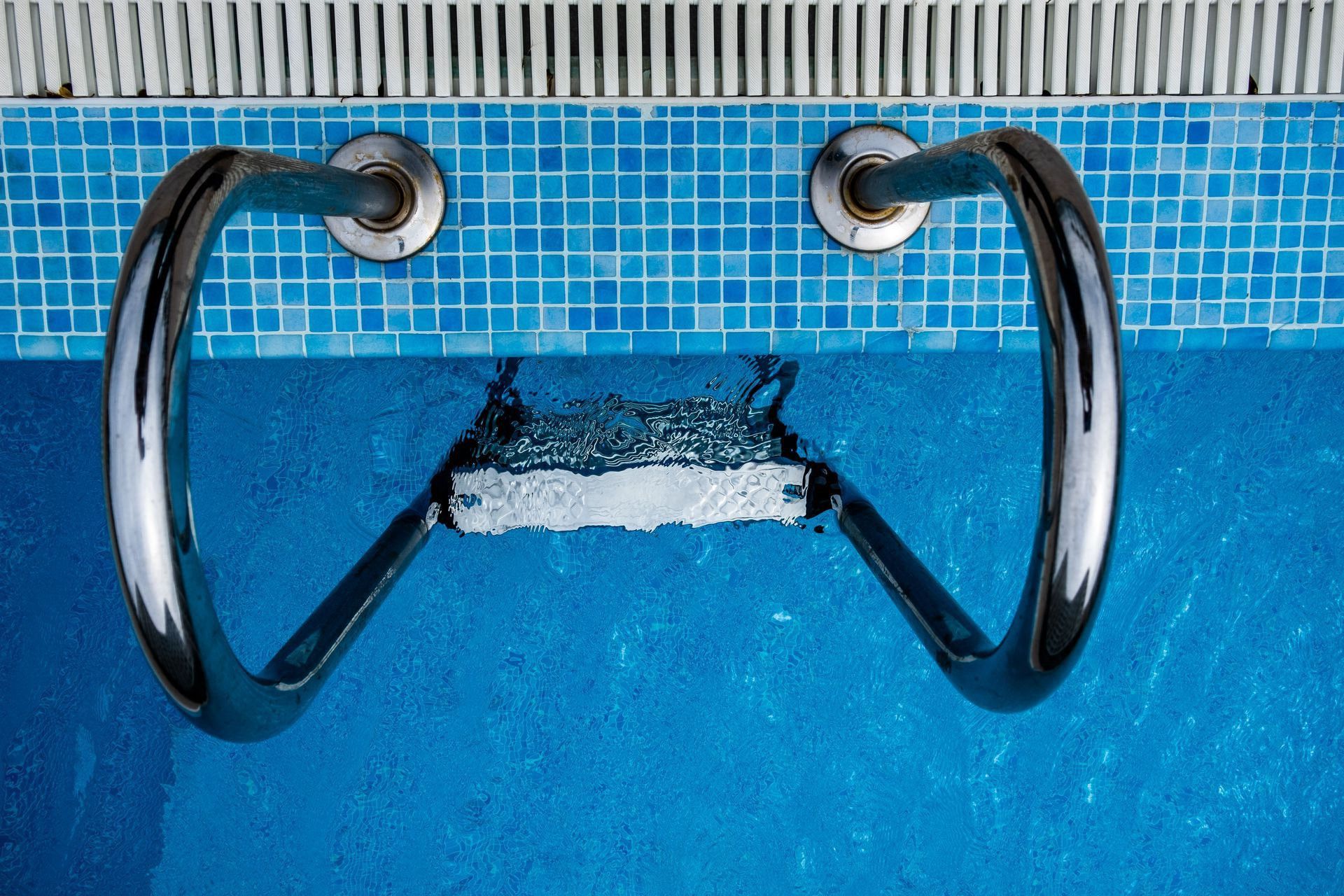 Aerial view of a stainless steel ladder entering a sparkling blue swimming pool with tiled edges.