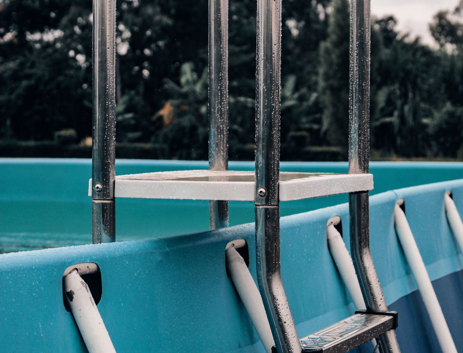 A detailed view of a metal ladder attached to an outdoor swimming pool, showcasing droplets and a serene nature backdrop.